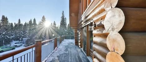 A wooden deck with snow-covered ground, a wooden railing, and a view of snow-covered trees and a car in the background.