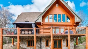 A log cabin with a stone foundation, a balcony with seating, and a large front door. - Sainte-Lucie Luxury, Chalet with Hot Tub, River & Sauna! (Sainte-Lucie-des-Laurentides)