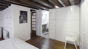 A bedroom with a wooden ceiling, a white bed, a white chair, and a door leading to a bathroom. - Peaceful appartement in the heart of Le Marais  (Paris)