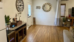 Un salón amplio con suelo de madera, un reloj de pared grande, un televisor y una puerta que da a otra habitación.