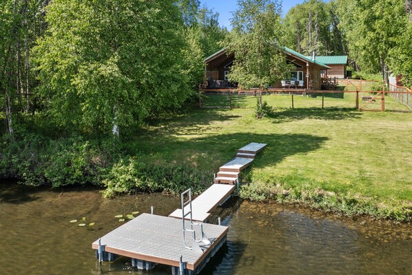 A wooden dock extending into a calm river, surrounded by lush greenery and a cabin with a green roof. - Wasilla 'Spruce Moose' Cabin: Lakefront + Hot Tub! (Wasilla)