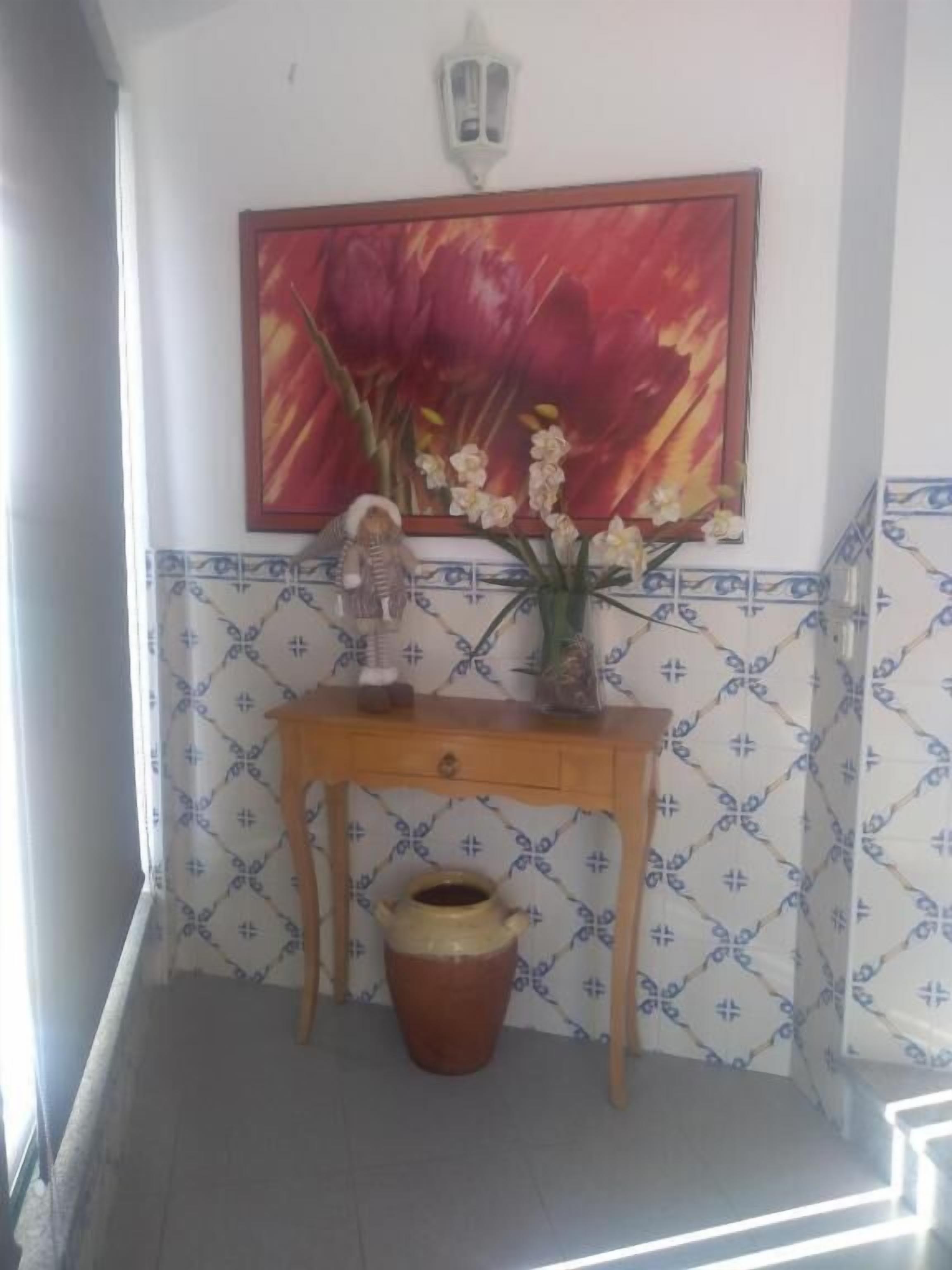 Una esquina con un muro de azulejos, una mesa de madera con un jarrón de flores, una maceta de cerámica y una lámpara fijada a la pared.