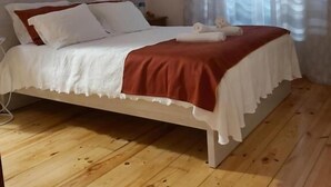 A bedroom with a wooden floor, a bed with white bedding and a red blanket, a nightstand, and a window with sheer curtains. - Encosta da Serra (Seia)