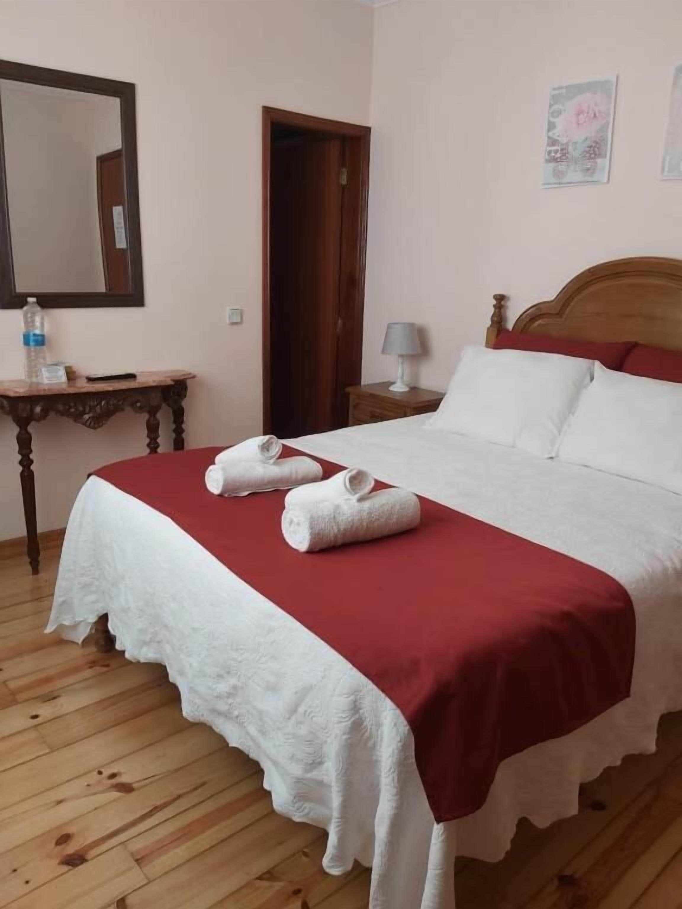 Una cama bien hecha con ropa de cama blanca y un detalle rojo, una mesita de noche de madera con una lámpara, una mesa de madera con una botella de agua y un espejo en la pared.