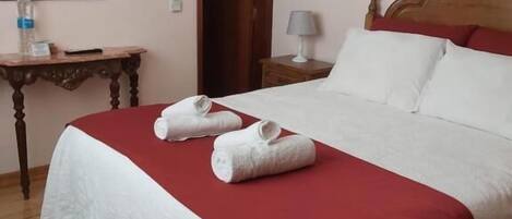 Una cama bien hecha con ropa de cama blanca y un detalle rojo, una mesita de noche de madera con una lámpara, una mesa de madera con una botella de agua y un espejo en la pared.