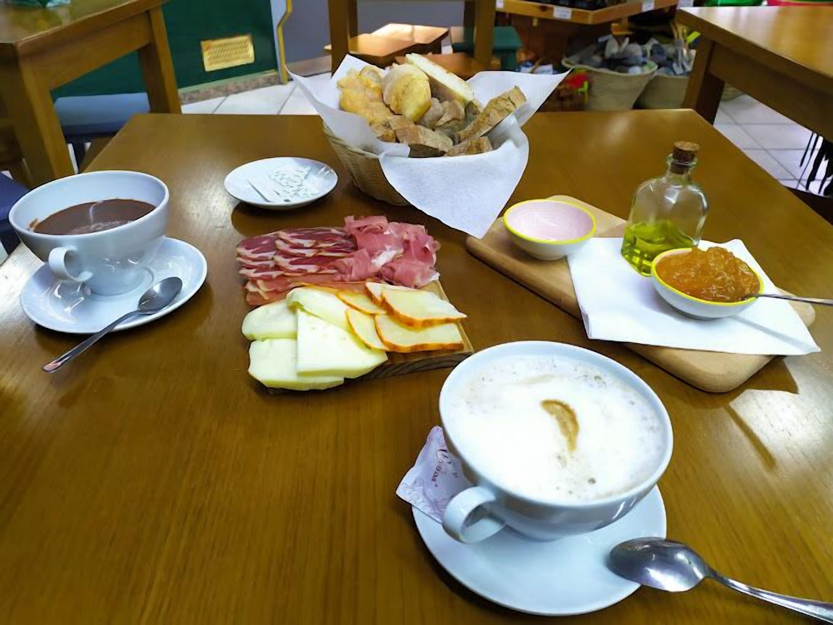 A wooden table with a breakfast setting including a cup of coffee, a bowl of soup, sliced cheese, cured meats, bread, and a bottle of sauce.
