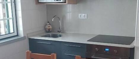 Cocina compacta con armarios oscuros, microondas, lavamanos y estufa. Hay una mesa de comedor de madera con sillas y una cesta de mimbre.