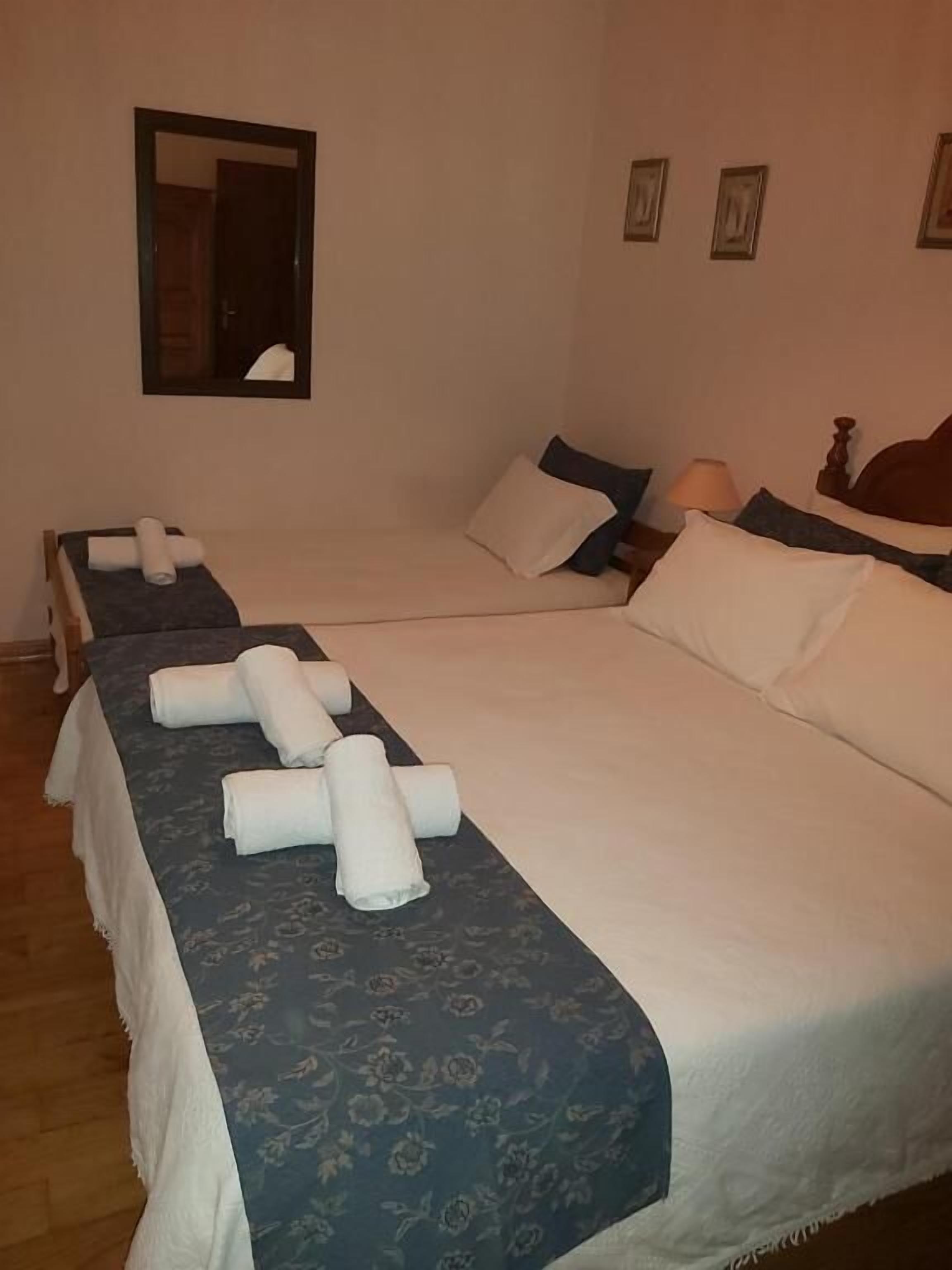 A hotel room with two beds, each with white linens and a blue floral bedspread. There is a wooden headboard on the right bed, a nightstand with a lamp, and a mirror on the wall.