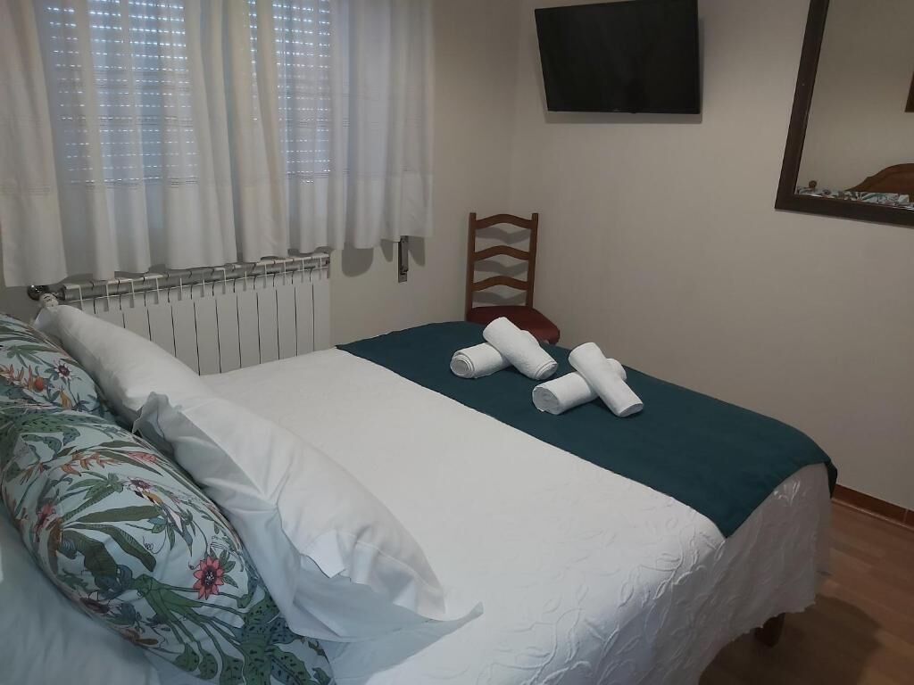 A neatly made bed with a teal bedspread and white pillows, a wooden chair, a radiator, and a flat-screen TV mounted on the wall.