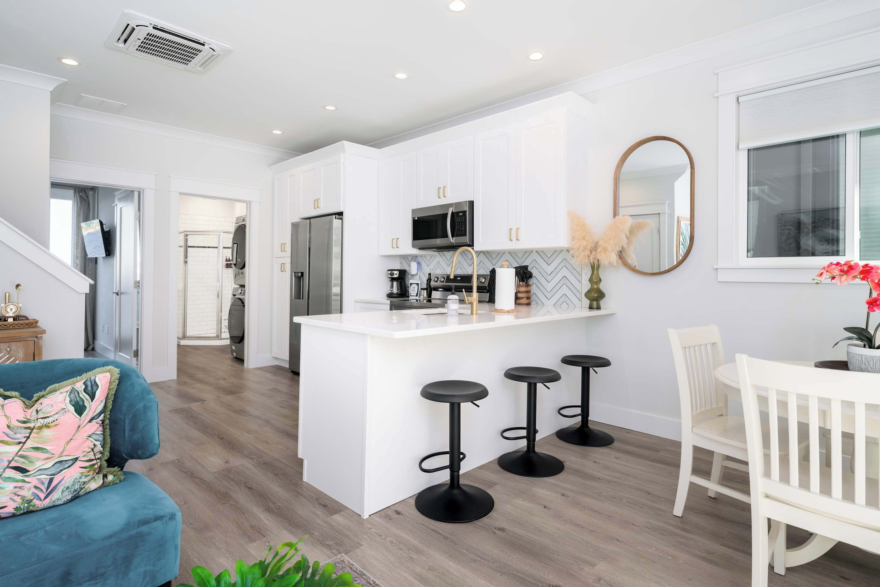 A modern kitchen with white cabinetry, a central island with stools, a dining area with a white table and chairs, and a large mirror on the wall.