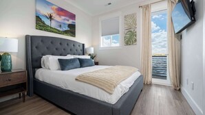 Ein Schlafzimmer mit einem großen Bett, einem Nachttisch, einem an der Wand montierten Fernseher und einem Fenster mit Vorhängen.