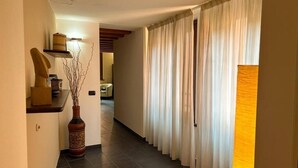 Un pasillo con piso de baldosas oscuras, un estante de madera con objetos decorativos, un jarrón alto con ramas y la entrada a una habitación con cortinas blancas.