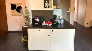 Una cocina con encimera negra, armarios blancos y una planta colgante. Hay varios utensilios de cocina sobre la encimera, como un hervidor, una cafetera y un cuenco rojo.