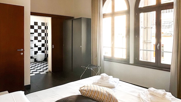 Una habitación de hotel con una cama grande, un armario, un baño con pared a cuadros y dos ventanas con cortinas.