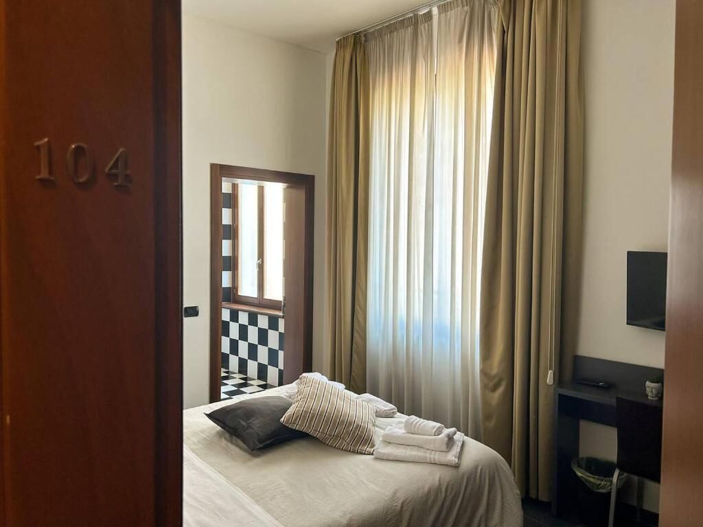 A hotel room with a bed, a checkered wall, a large window with curtains, and a TV on a stand.