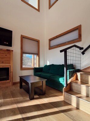 A modern living room with a green sofa, a wooden coffee table, and a fireplace. There are windows with blinds and a staircase with a railing.
