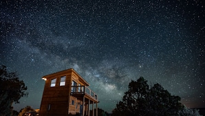 Una cabaña de madera con balcón, rodeada de árboles, bajo un cielo estrellado.