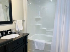 Un baño con tocador negro, lavamanos blanco, espejo y zona de ducha con mampara de cristal que incluye una bañera blanca.