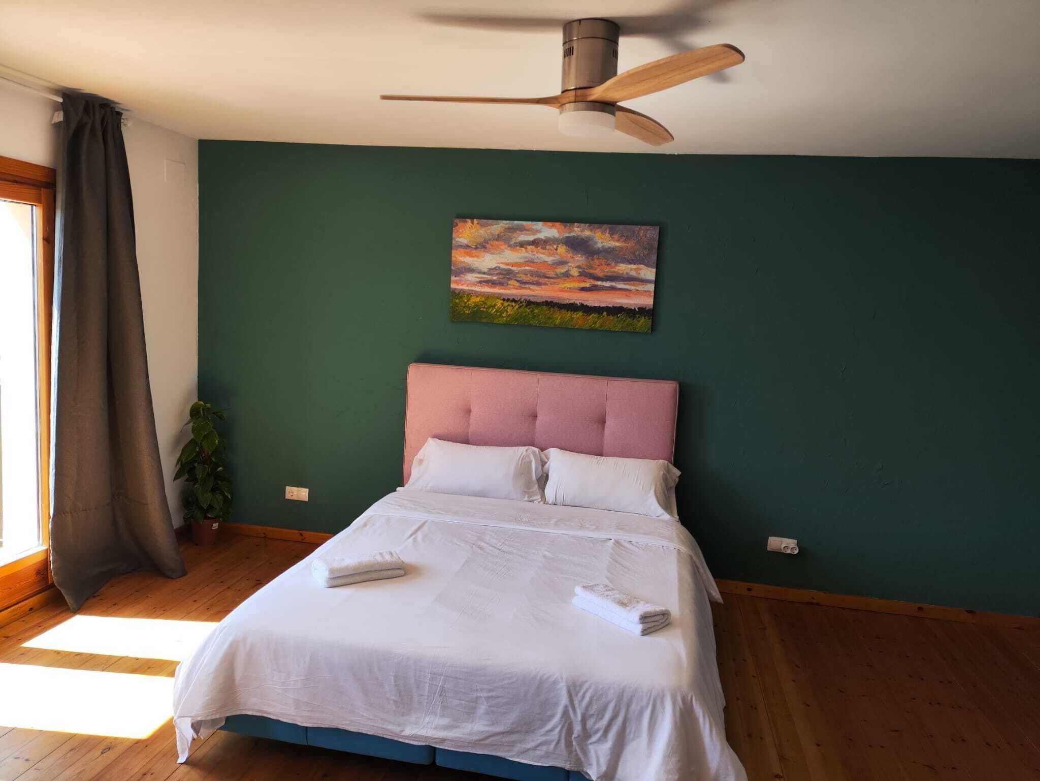Un dormitorio con cama, ventilador de techo, un cuadro y ventana con cortinas.