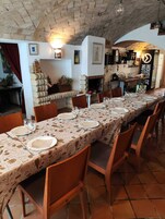 Una larga mesa de comedor dispuesta con platos, vasos y servilletas, rodeada de sillas de madera en una habitación rústica con paredes de ladrillo visto y una chimenea de ladrillo.