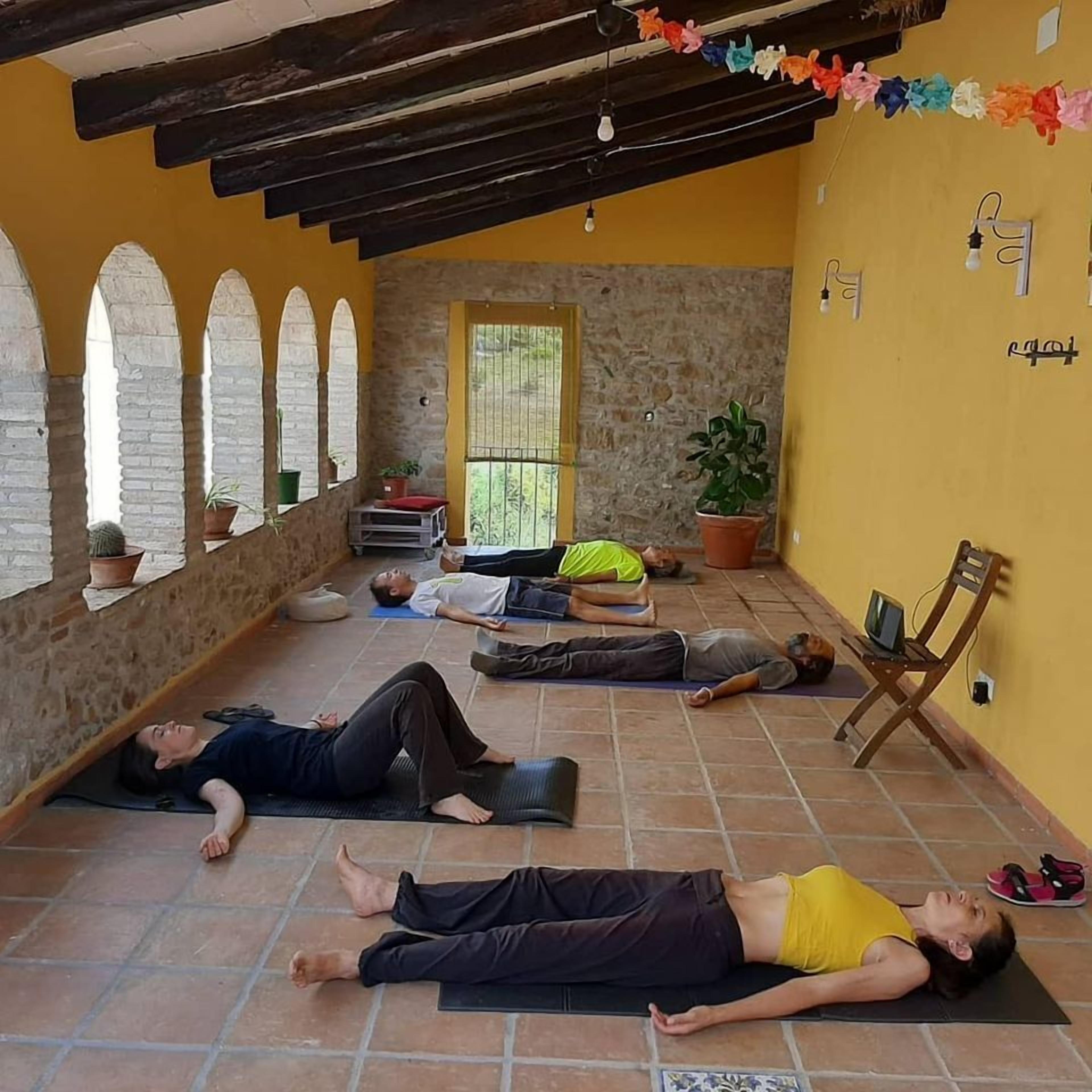 Una habitación con pared de piedra, techo de madera y pared amarilla con ventana. Cuatro personas descansan sobre esterillas en el suelo.