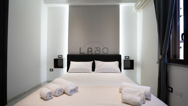 A hotel room with a bed, two bedside tables, a wall-mounted air conditioner, and a window with curtains. - Ammos Guest House (Tropea)