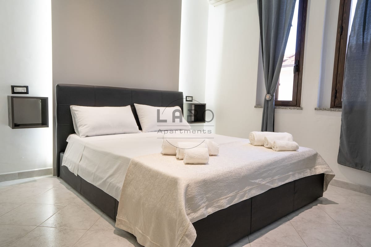 A neatly made bed with a dark headboard and white linens, a bedside table with a lamp, and a window with curtains.