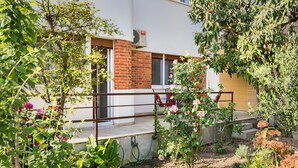 Un edificio residenziale con facciata in mattoni, un balcone con sedie rosse e un giardino con diverse piante e fiori.