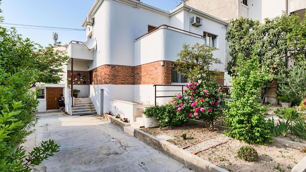 Un edificio residenziale a due piani con facciata bianca e dettagli in mattoni, un ingresso con una scalinata e un giardino con piante in fiore.