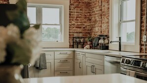 A kitchen with exposed brick walls, white cabinetry, and a large window. - Charming renovated guesthaus in the heart of Historic Hermann. (Hermann)