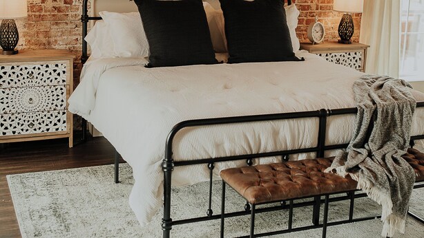 A bedroom with a brick wall, a bed with black metal frame, white bedding, two bedside tables with lamps, a wooden bench, and a floral artwork on the wall. - Charming renovated guesthaus in the heart of Historic Hermann. (Hermann)