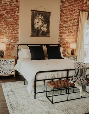 Une chambre avec un mur en briques, un lit avec un cadre en métal noir, une literie blanche, deux tables de chevet avec des lampes, un banc en bois et une œuvre d’art florale accrochée au mur.