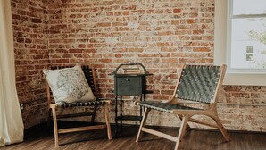 A room with a brick wall, two wooden chairs with cushions, a small wooden table, and a window with a view of greenery. - Charming renovated guesthaus in the heart of Historic Hermann. (Hermann)
