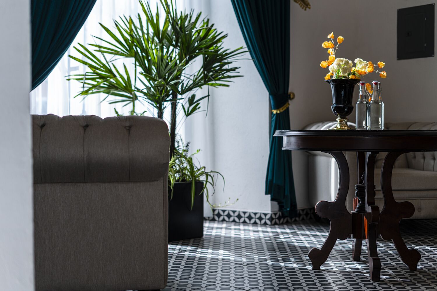 Una habitación con un sofá capitonado, una mesa con un jarrón de flores, una planta en maceta y un suelo estampado.