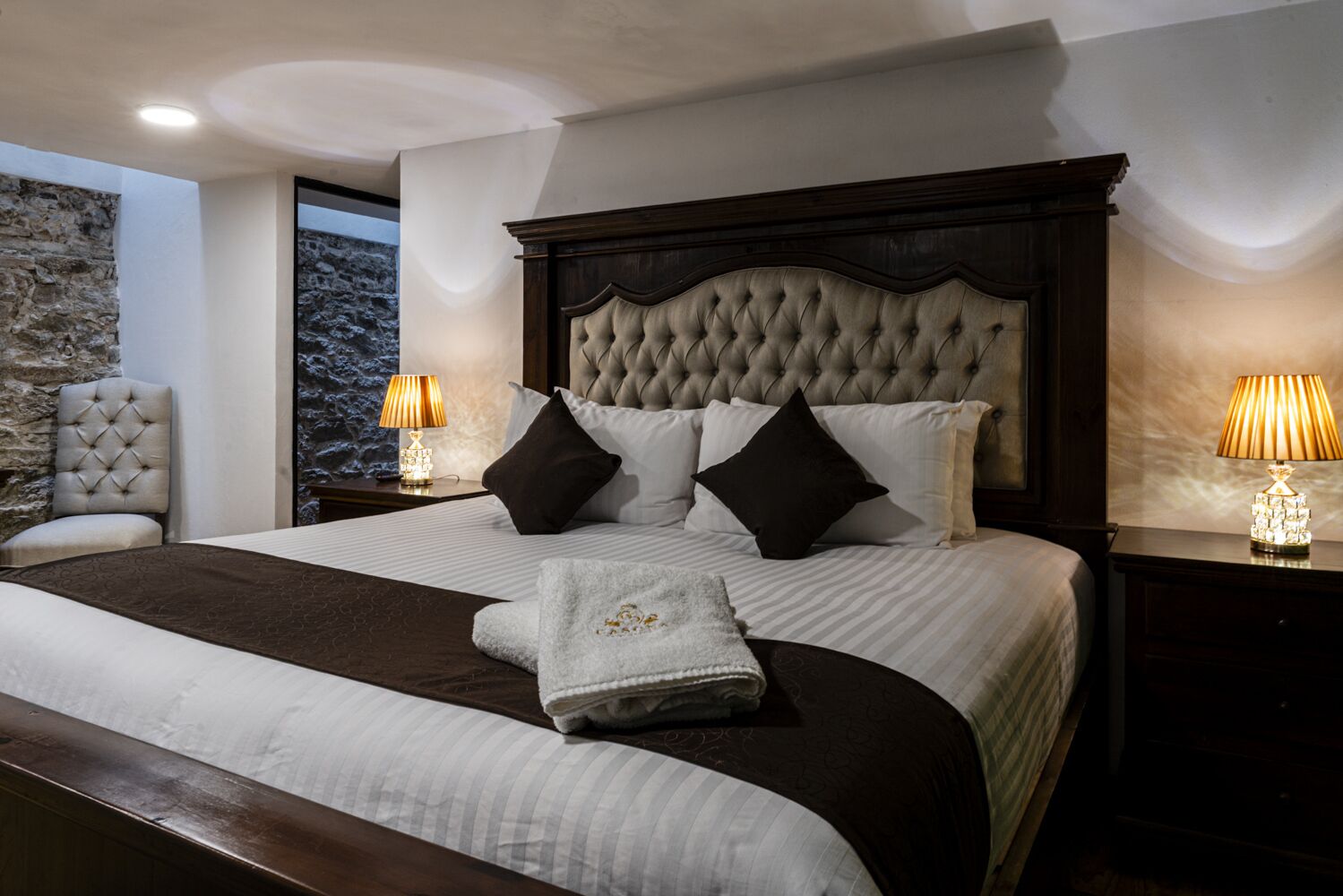 A large bed with a tufted headboard, two bedside tables with lamps, a chair, and a stone wall.
