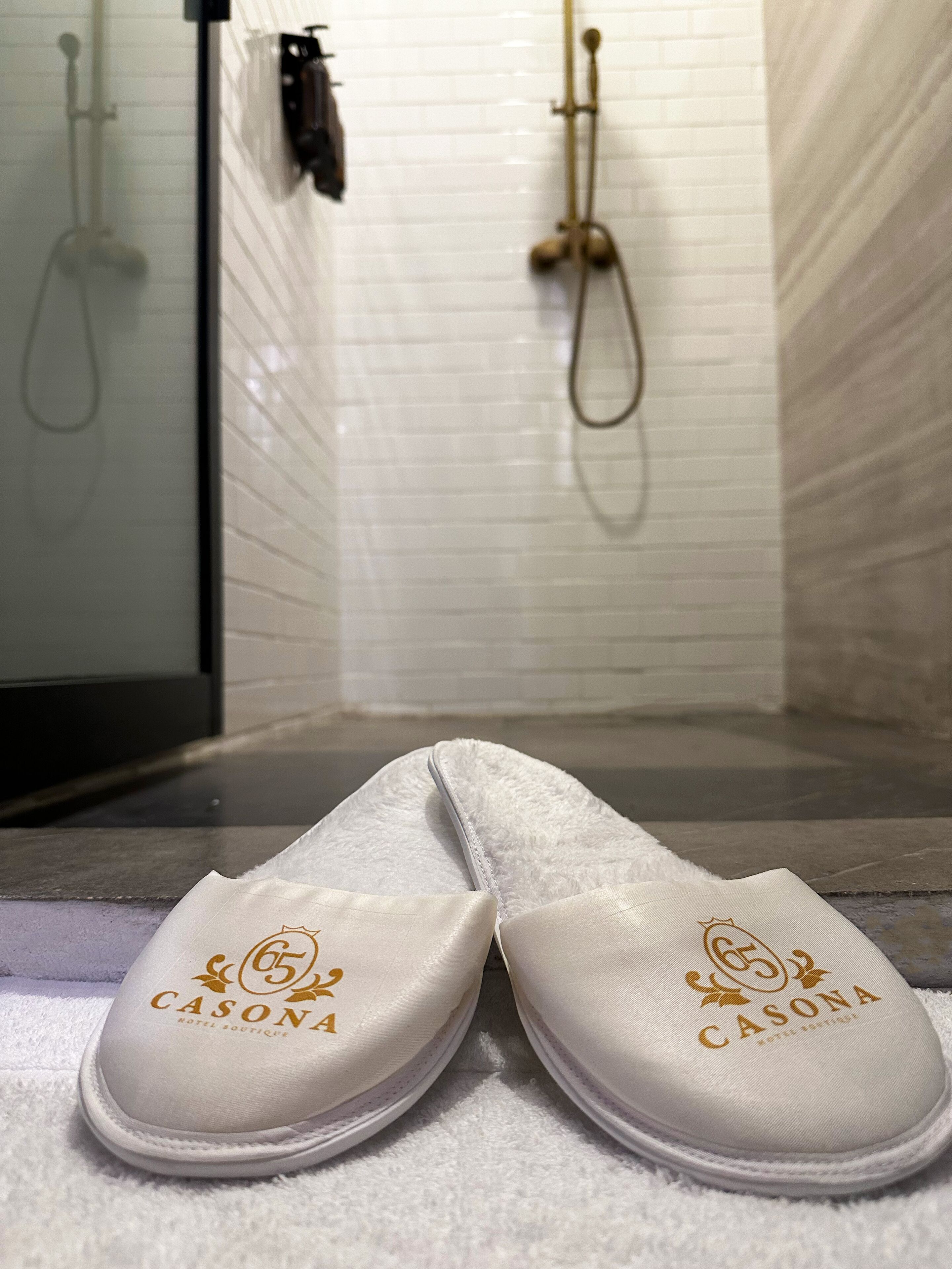 A pair of white slip-on shoes with a logo on the sole, placed on a bathroom floor.