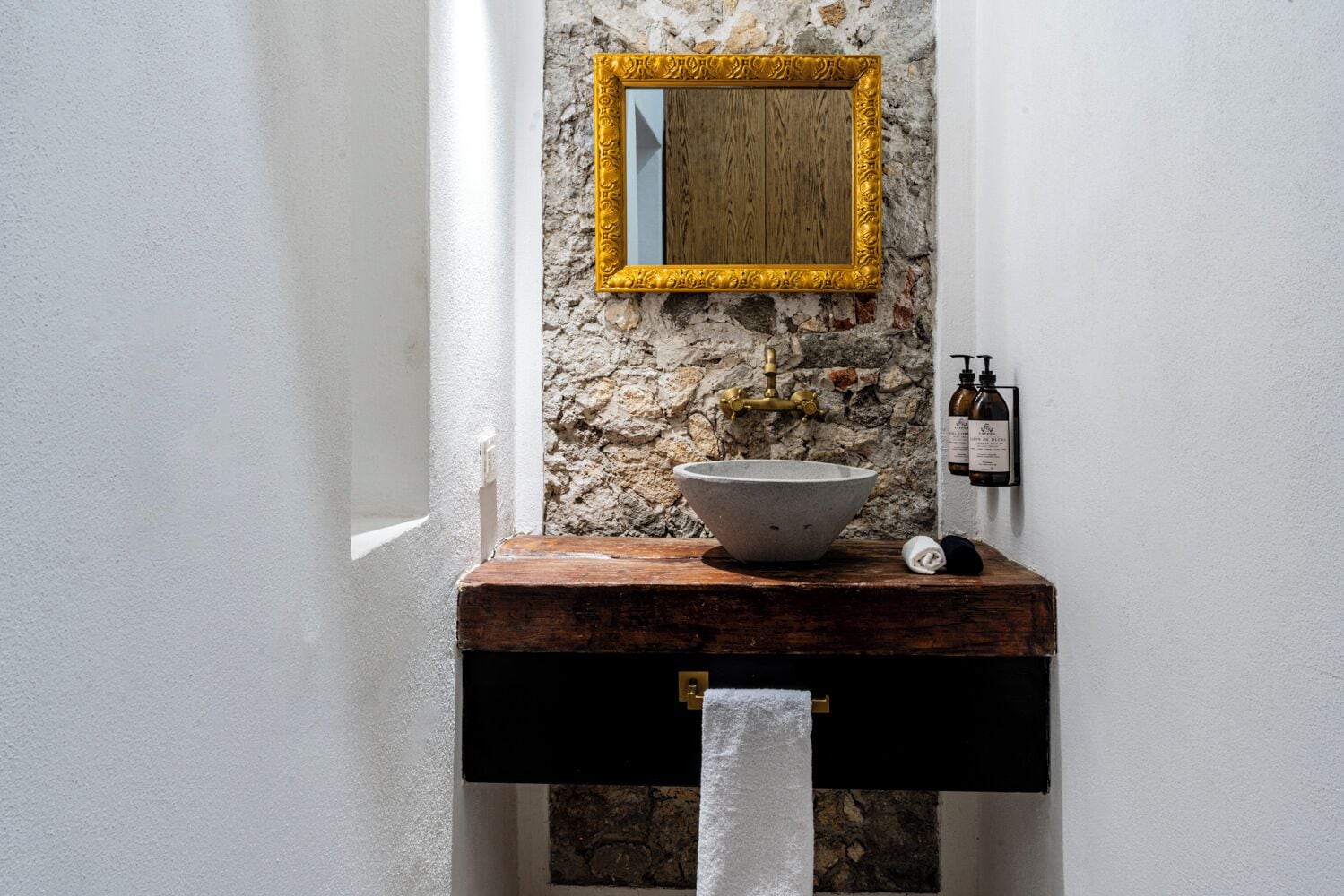 A bathroom with a stone sink, a wooden vanity, a gold-framed mirror, and a towel rack.