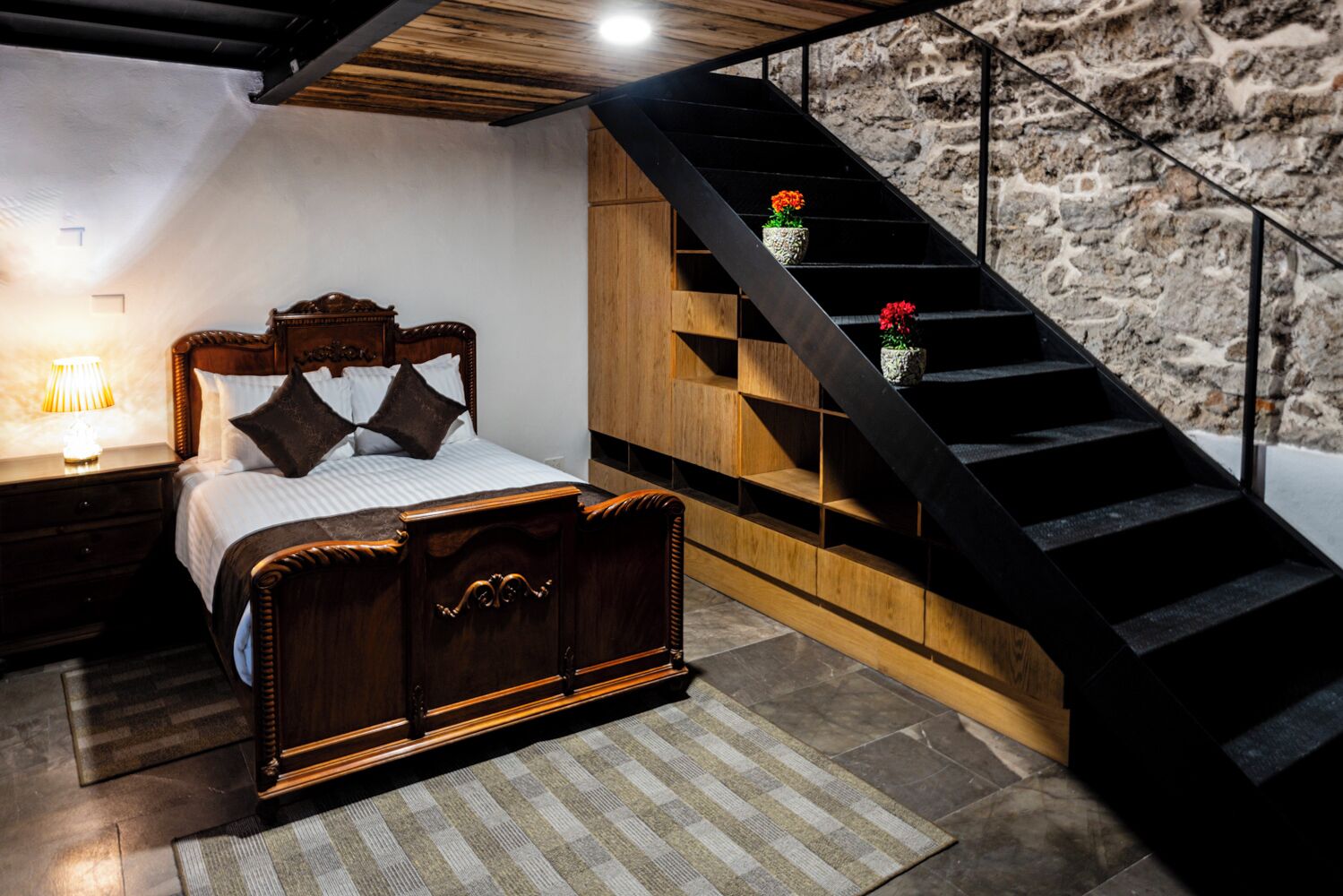 A bedroom with a wooden bed, a nightstand, a lamp, a staircase with a stone wall, and a small potted plant.