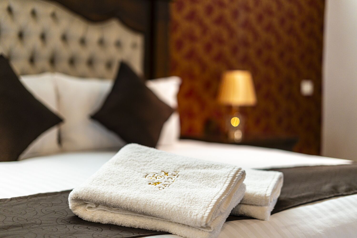 A neatly made bed with white and dark gray linens, a folded white towel with a decorative pattern, and a lamp in the background.