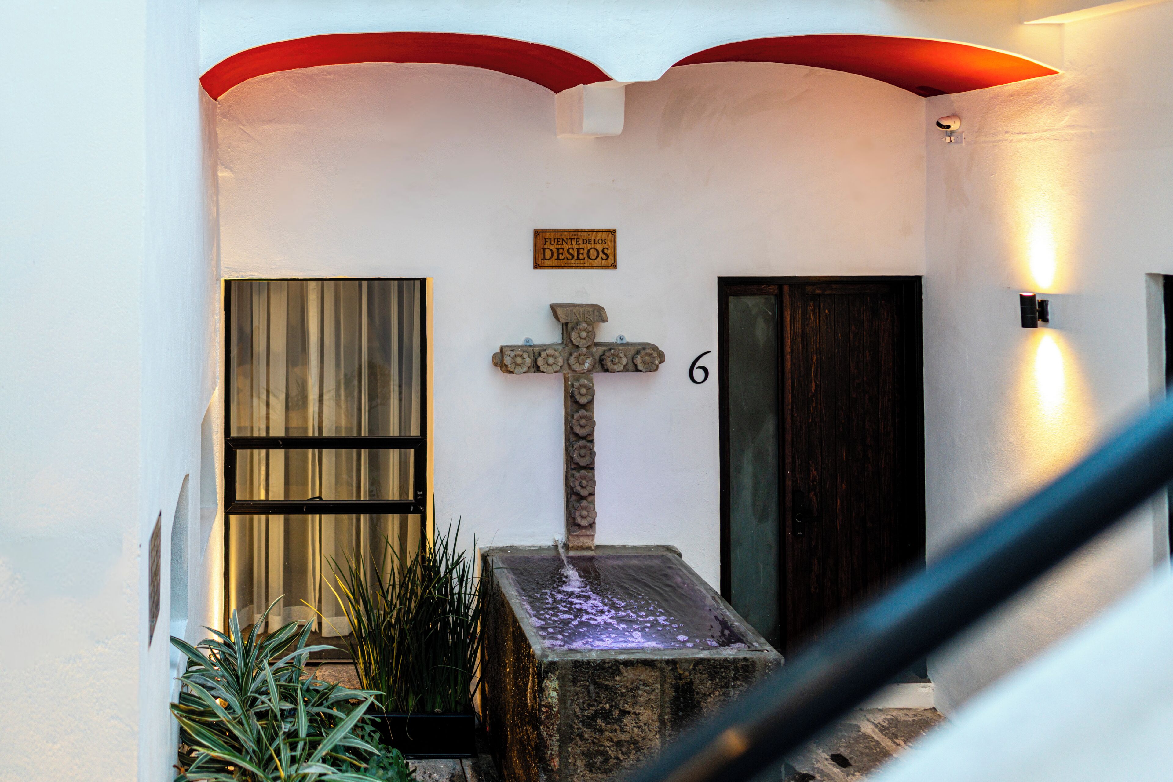 A stone cross with a water feature in front of a building with a sign reading "Fuente de Deseos" and the number 6.