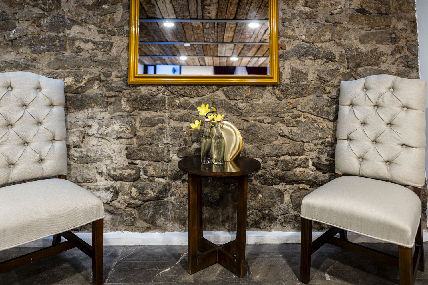 Two upholstered chairs with a tufted design, a small round wooden table with a vase of flowers, and a mirror on a stone wall.