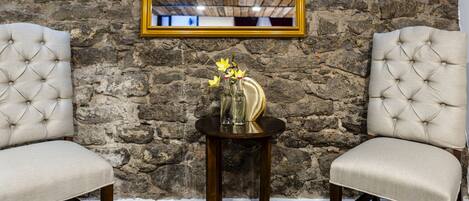 Two upholstered chairs with a tufted design, a small round wooden table with a vase of flowers, and a mirror on a stone wall.