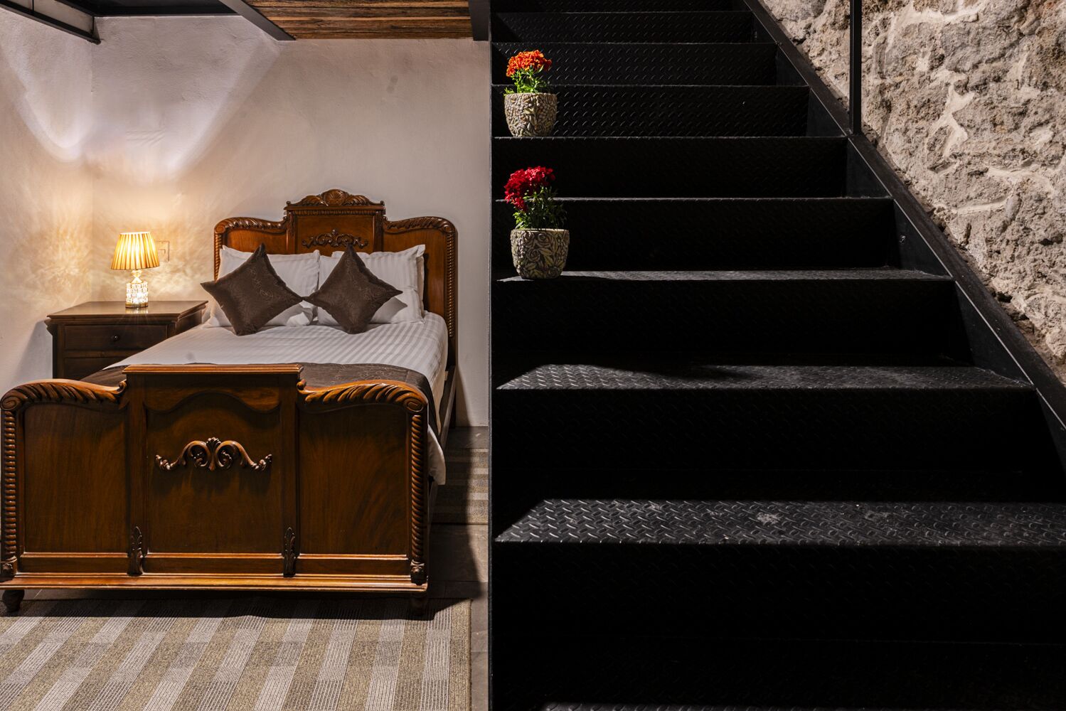 A bedroom with a wooden bed, a nightstand with a lamp, and a staircase with potted plants.