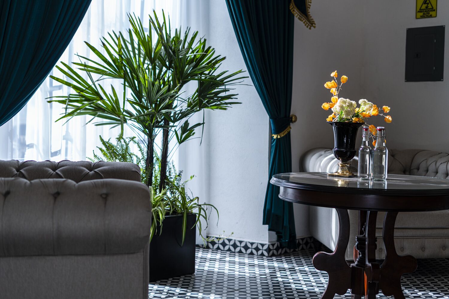 Una habitación con un sofá capitonado, una planta en maceta, una mesa redonda con un jarrón de flores y un suelo estampado.