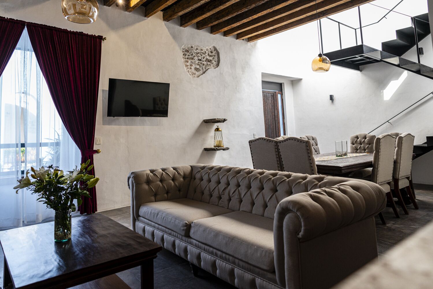 Una sala de estar moderna con un sofá capitonado, una mesa de comedor con sillas, un televisor montado en la pared y una escalera que sube a un nivel superior.