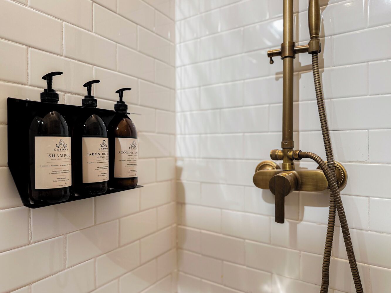 Un baño con paredes de azulejos blancos, un estante para dispensador de jabón negro y un cabezal de ducha de latón.