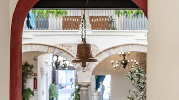 Une cloche suspendue dans un portail en arc, d’où l’on aperçoit un couloir et un balcon orné de plantes.