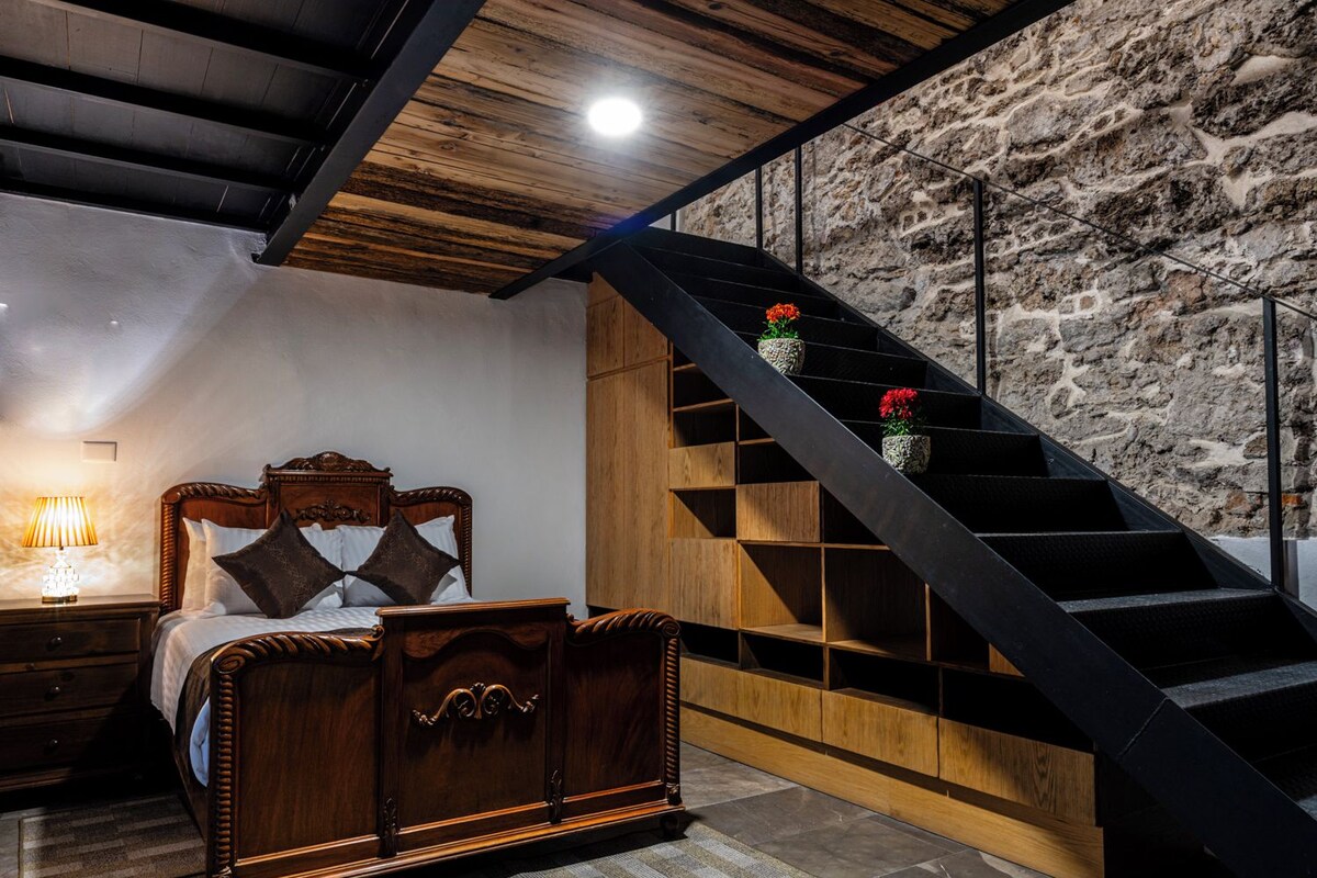Un dormitorio con cama de madera, mesita de noche, lámpara, pared de piedra y escalera adornada con plantas en macetas.