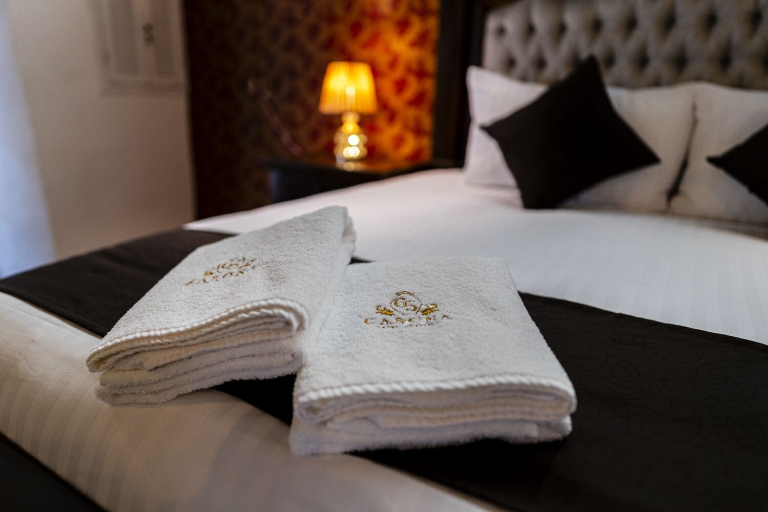 A neatly made bed with white and black bedding, two folded towels with embroidered details, a lamp, and a patterned wall in the background.