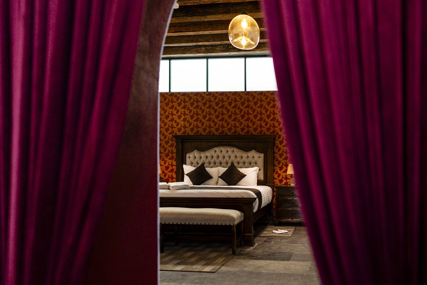 A bedroom with a tufted headboard, a wooden nightstand, and a patterned wallpaper.